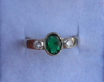 May include: A silver ring featuring an oval-cut, emerald-green gemstone in the center, flanked by two smaller, round, clear gemstones. The ring is set against a light blue background.