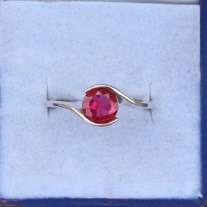 May include: A silver ring with a large, round, bright red gemstone. The band curves around the gemstone, creating a modern and elegant design. The ring is set against a light blue background.