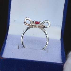 May include: A silver ring with a bow-shaped design. The bow features a central red gemstone and is adorned with small, clear stones. The ring is presented in a blue jewelry box with a white interior, creating a striking contrast.