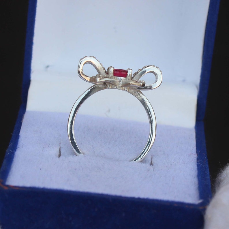 May include: A silver ring with a bow-shaped design. The bow features a central red gemstone and is adorned with small, clear stones. The ring is presented in a blue jewelry box with a white interior, creating a striking contrast.