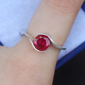 May include: A silver ring featuring a large, round, vibrant red gemstone. The band curves around the stone in a flowing, modern design. The ring is shown on a finger, highlighting the gemstone's color and setting.