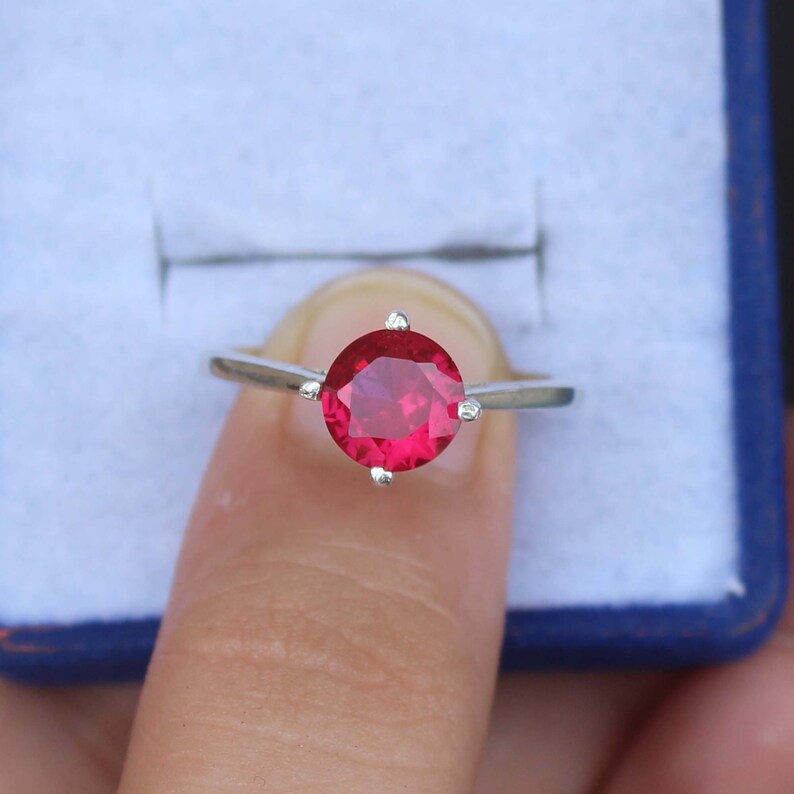K&ouml;nnte beinhalten: Ein silberner Ring mit einem gro&szlig;en, runden, leuchtend roten Edelstein. Der Edelstein wird von vier kleinen Krappen gehalten. Der Ring wird vor einem hellblauen Hintergrund gezeigt.