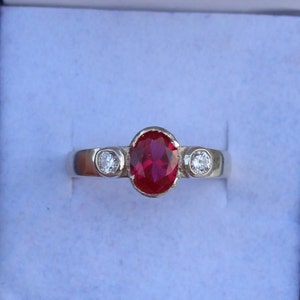 May include: A silver ring with a central oval-cut ruby and two smaller, round, clear gemstones. The ring is set against a light blue background. The gemstones are set in a silver band.