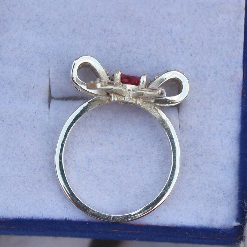 May include: A silver-toned ring with a bow-shaped design. The bow features a rectangular, red gemstone at its center and is accented with small, clear stones. The ring is set against a light blue background.