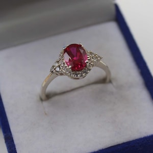May include: A silver ring with a large oval, ruby-red gemstone at the center, surrounded by smaller, clear stones. The ring is set in a blue box, suggesting a jewelry presentation.