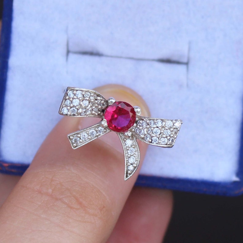 May include: A silver-toned bow ring with a large, round, red gemstone at the center. The bow is encrusted with small, clear gemstones. The ring is held in a blue jewelry box.