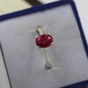 May include: A silver ring featuring a vibrant oval-cut ruby gemstone. The ruby is a deep red color and is held in place by four prongs. The ring is set against a blue background.