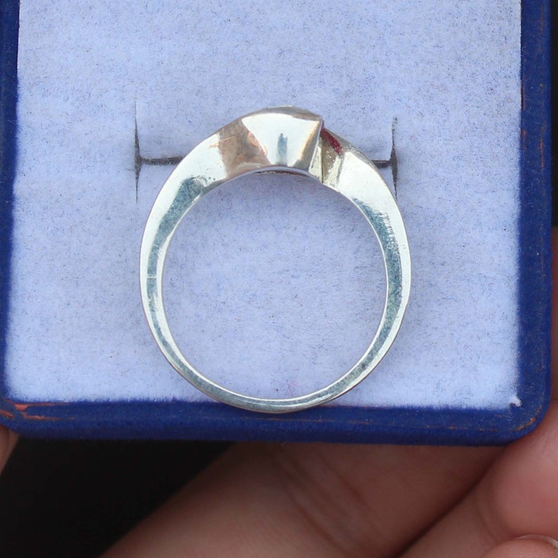 May include: A silver ring with a geometric design, featuring a square-cut gemstone. The ring is set against a blue background, showcasing its polished metal and the clear facets of the stone. A simple, elegant piece of jewelry.