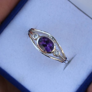 Puede incluir: Un anillo de plata con una piedra ovalada púrpura y dos piedras redondas blancas más pequeñas. El anillo tiene una banda texturizada.