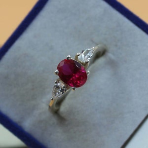 Puede incluir: Un anillo de plata con una gran piedra de rubí de forma ovalada y dos piedras blancas más pequeñas en forma de pera.
