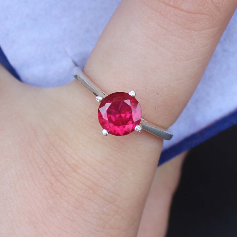 K&ouml;nnte beinhalten: Ein silberner Ring mit einem gro&szlig;en, runden, leuchtend roten Edelstein. Der Edelstein wird von vier kleinen Krappen gehalten. Der Ring hat ein schlichtes, poliertes Silberband.
