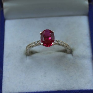 May include: A silver ring with a large oval-cut ruby gemstone as the centerpiece. The band is adorned with small, sparkling accent stones. The ring is presented in a blue jewelry box, highlighting its elegant design.
