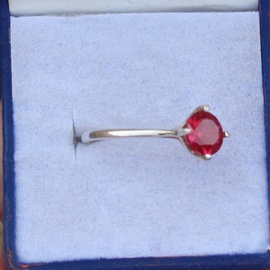 K&ouml;nnte beinhalten: Ein silberner Ring mit einem gro&szlig;en, runden, rubinroten Edelstein. Der Ring hat eine Vier-Zacken-Fassung und ein d&uuml;nnes, poliertes Band. Der Ring wird in einer blauen Schmuckschatulle pr&auml;sentiert.