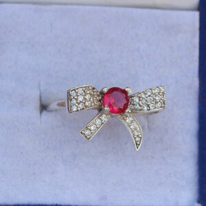 May include: A silver ring featuring a bow design. The bow is encrusted with small, clear gemstones and a large, round, red gemstone at the center. The ring is set against a light blue background.