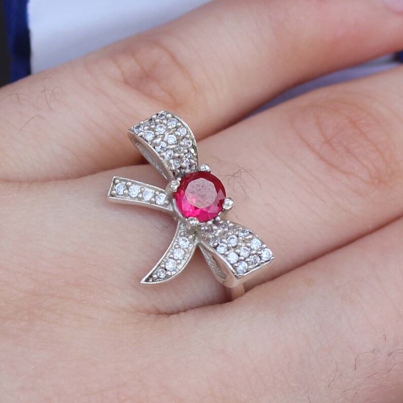 May include: A silver ring in a bow design, adorned with clear, sparkling stones and a central, round, red gemstone. The ring is shown on a person's finger, highlighting the intricate details of the jewelry.