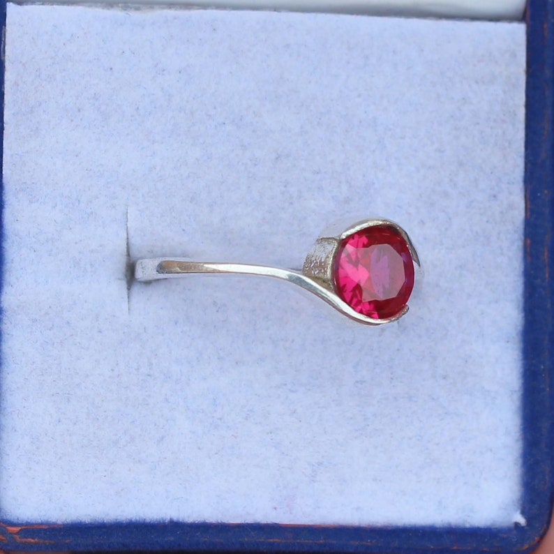 May include: A silver ring featuring a large, round, vibrant red gemstone. The gemstone is set in a bezel setting, and the band is a simple, elegant design. The ring is displayed in a blue jewelry box.