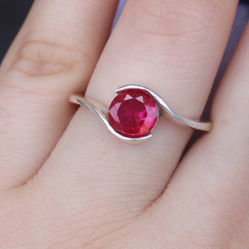May include: A silver ring featuring a large, round, vibrant red gemstone. The band curves around the stone, creating a modern, elegant design. The gemstone is the focal point, catching the light and drawing the eye.