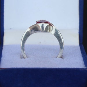 May include: A silver ring with a red gemstone, presented in a blue jewelry box. The ring's band curves gracefully, leading to the prominent, vibrant red stone. The setting appears to be a simple, yet elegant design.