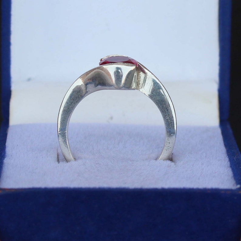 May include: A silver ring with a red gemstone, presented in a blue jewelry box. The ring's band curves gracefully, leading to the prominent, vibrant red stone. The setting appears to be a simple, yet elegant design.