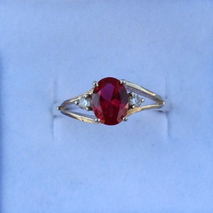 Könnte beinhalten: Ein silberner Ring mit einem großen, oval geschliffenen, tiefroten Edelstein als Mittelstück. Zwei kleinere, runde, klare Edelsteine flankieren den Hauptstein. Der Ring hat ein geteiltes Schaftdesign.