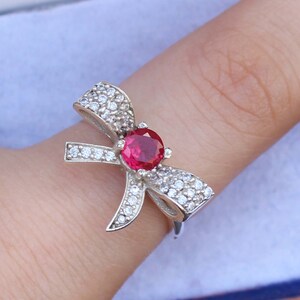 May include: A silver ring in a bow shape, adorned with small, clear gemstones and a large, round, red gemstone at the center. The ring is on a finger, showcasing the design and sparkle of the jewelry.