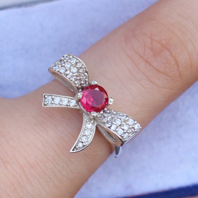 May include: A silver ring in a bow shape, adorned with small, clear gemstones and a large, round, red gemstone at the center. The ring is on a finger, showcasing the design and sparkle of the jewelry.