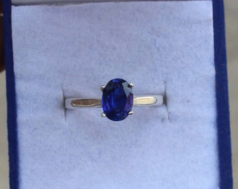 Op de afbeelding: Een zilveren ring met een grote, ovaal geslepen, diepblauwe edelsteen. De ring is gezet in een eenvoudige vier-klauwzetting en wordt gepresenteerd in een blauwe ringdoos. De edelsteen is het middelpunt en toont zijn rijke kleur en schittering.