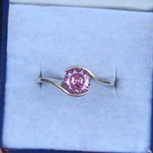 Può includere: Un anello d'argento con una pietra preziosa di colore viola chiaro incastonata in una fascia curva.