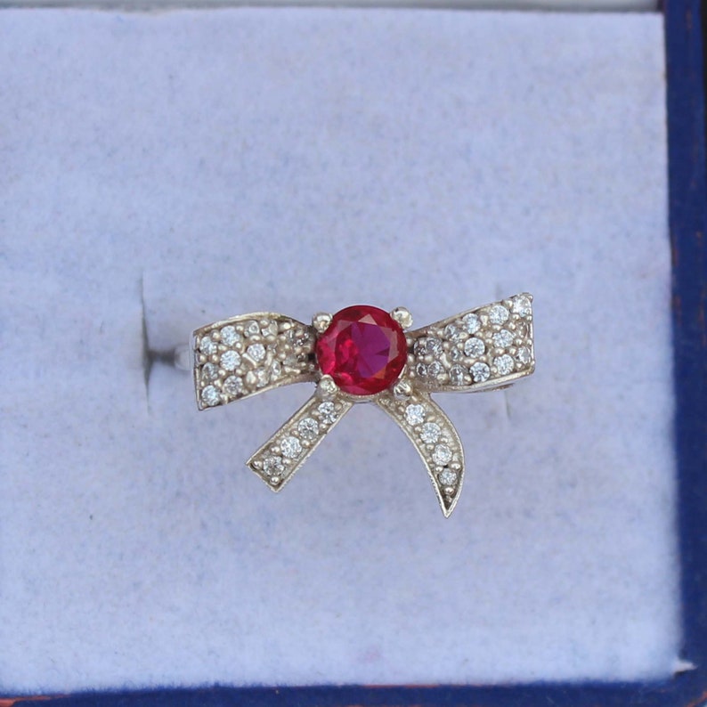 May include: A silver bow ring with a large, round, red gemstone at the center. The bow is encrusted with small, clear gemstones. The ring is set against a light blue background.