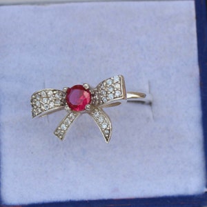 May include: A silver-toned ring featuring a bow design. The center of the bow holds a round, red gemstone, surrounded by smaller, clear gemstones. The ring is set against a light blue background.