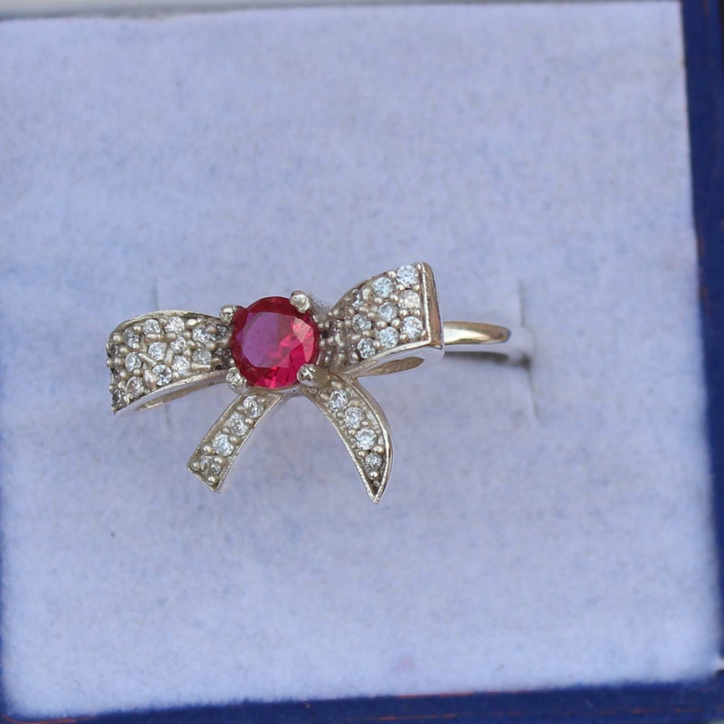 May include: A silver-toned ring featuring a bow design. The center of the bow holds a round, red gemstone, surrounded by smaller, clear gemstones. The ring is set against a light blue background.