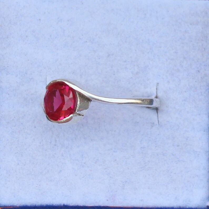 May include: A silver ring featuring a vibrant red oval gemstone. The gemstone is the focal point, set in a sleek, minimalist design. The ring is displayed against a light blue background.