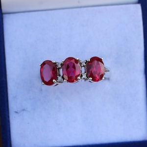 Op de afbeelding: Een zilveren ring met drie ovale rode edelstenen en kleinere heldere edelstenen ertussen.