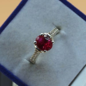 May include: A silver ring with a large, oval-shaped ruby gemstone surrounded by smaller, clear gemstones.