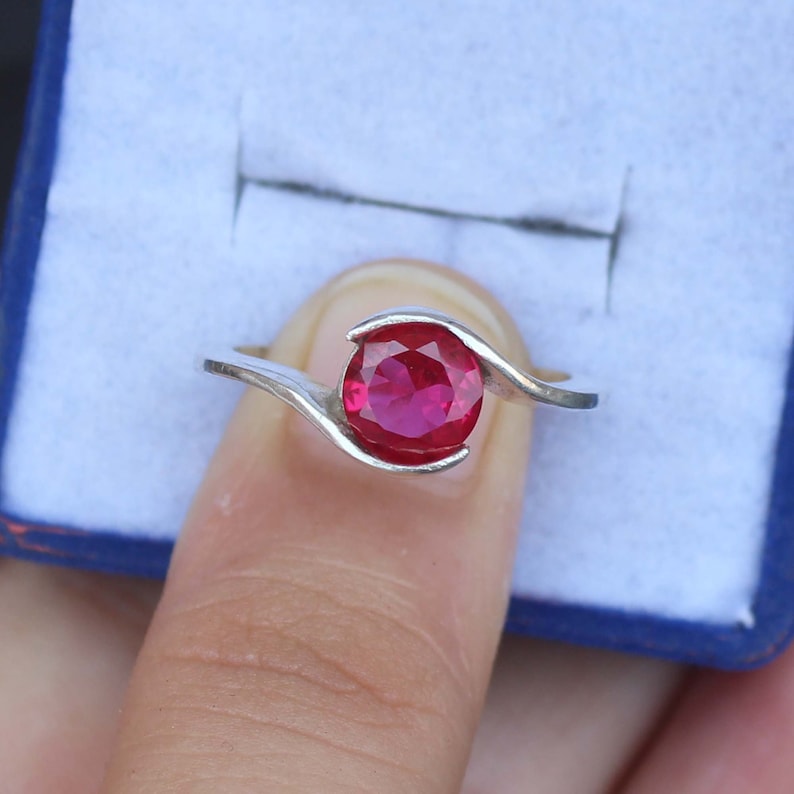 May include: A silver ring featuring a vibrant, round, red gemstone. The gemstone is the focal point, set in a swirling, minimalist design. The ring is displayed against a blue background.