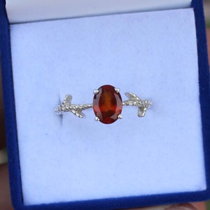 May include: A silver ring with a brown oval gemstone set in the center. The ring band is designed to look like a branch with two leaves.
