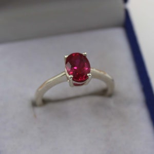 Puede incluir: Un anillo de plata con una vibrante gema de rub&iacute; de corte ovalado. El rub&iacute; es de un color rojo intenso y est&aacute; engastado en un engaste cl&aacute;sico de cuatro puntas. La banda es de plata pulida sencilla. El anillo se muestra en una caja de joyer&iacute;a azul.