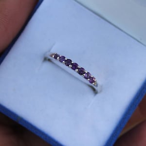 Könnte beinhalten: Ein silberner Ring mit einer Reihe kleiner, runder, violetter Edelsteine. Der Ring befindet sich in einer hellblauen Schachtel, was darauf hindeutet, dass es sich um ein Schmuckstück handelt. Die Edelsteine sind gleichmäßig angeordnet.