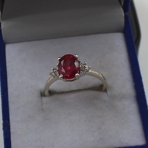Pode incluir: Anel de prata com um grande rubi oval de cor vermelha intensa. O rubi é ladeado por pedras preciosas menores e transparentes de cada lado. O anel, com um design clássico, é apresentado numa caixa azul, ideal para ocasiões especiais.