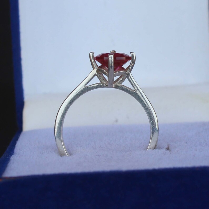 K&ouml;nnte beinhalten: Ein silberner Ring mit einem gro&szlig;en, facettierten, roten Edelstein. Der Edelstein wird von vier Krappen gehalten. Der Ring liegt auf blauem Samt in einer Schmuckschatulle.