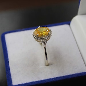 Op de afbeelding: Een zilveren ring met een grote gele ovale edelsteen omringd door kleinere heldere edelstenen.