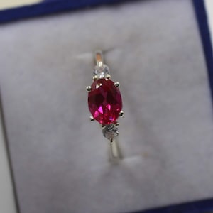 May include: A silver ring with a large oval-shaped ruby gemstone and two smaller round white gemstones on either side.