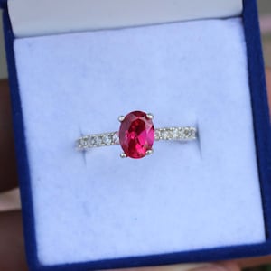 May include: A silver ring with a large oval-cut, vibrant red gemstone as the focal point. The band is adorned with smaller, clear gemstones. The ring is presented in a blue jewelry box, showcasing its elegant design.