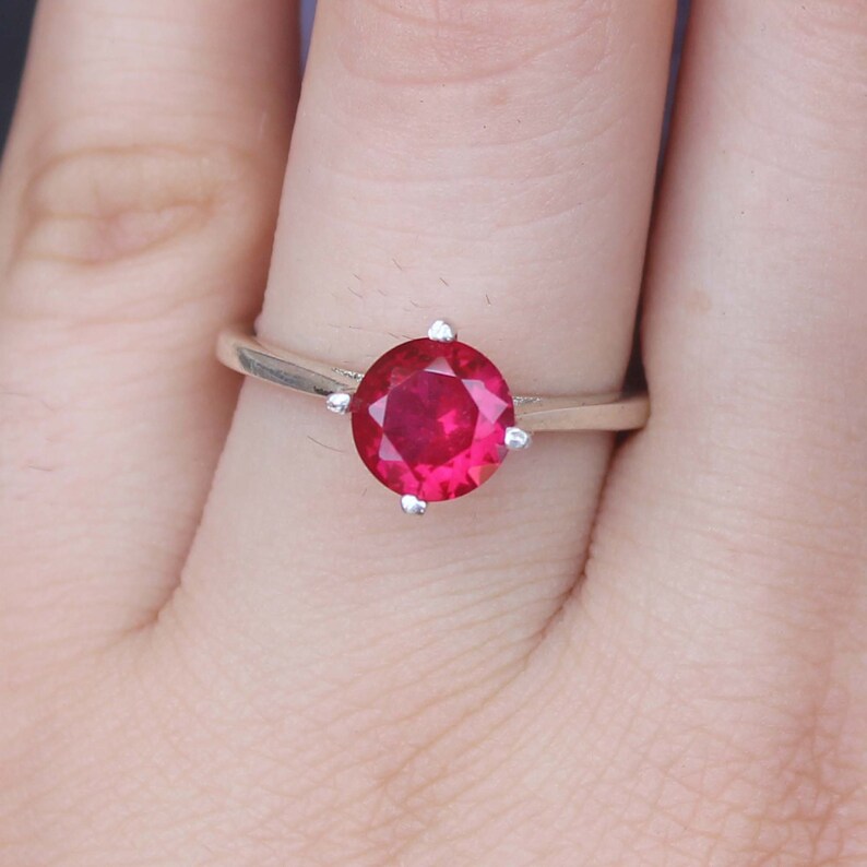 K&ouml;nnte beinhalten: Ein silberner Ring mit einem gro&szlig;en, runden, leuchtend roten Edelstein. Der Edelstein wird von vier kleinen Krappen gehalten. Der Ring wird an einem Finger gezeigt, wodurch die Farbe und die Fassung des Edelsteins hervorgehoben werden.