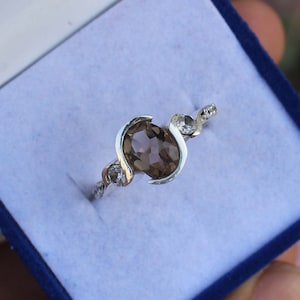 May include: A silver ring featuring a large, oval, smoky brown gemstone. The ring has a unique, flowing design with smaller, clear gemstones set along the band. The ring is displayed in a blue jewelry box.