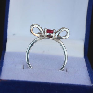 May include: A silver ring with a bow design, featuring a rectangular red gemstone at the center. The bow's loops are adorned with small, clear stones. The ring is presented in a blue jewelry box.