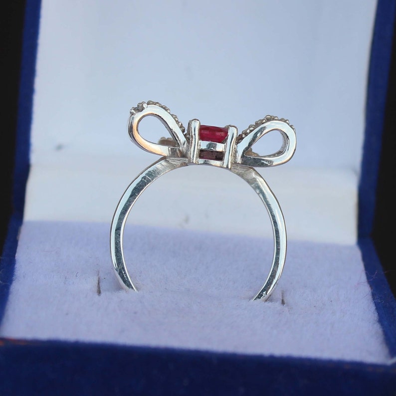 May include: A silver ring with a bow design, featuring a rectangular red gemstone at the center. The bow's loops are adorned with small, clear stones. The ring is presented in a blue jewelry box.