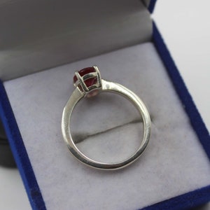 Puede incluir: Un anillo de plata con una gema redonda de color rojo intenso engastada en un engaste de cuatro puntas. El anillo se muestra dentro de una caja de joyer&iacute;a azul con interior blanco. La banda del anillo es lisa y pulida.