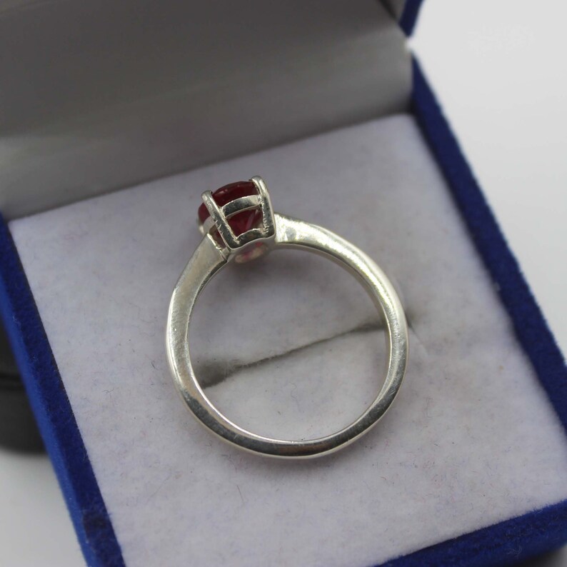 Puede incluir: Un anillo de plata con una gema redonda de color rojo intenso engastada en un engaste de cuatro puntas. El anillo se muestra dentro de una caja de joyer&iacute;a azul con interior blanco. La banda del anillo es lisa y pulida.