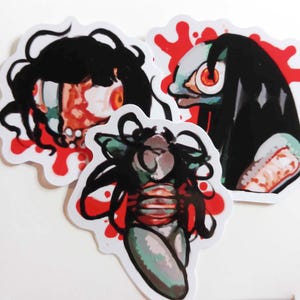 Puede incluir: Tres pegatinas con ilustraciones de mujeres con cabello negro largo y salpicaduras de sangre roja y blanca. Las mujeres tienen ojos grandes y están dibujadas en un estilo de dibujos animados.